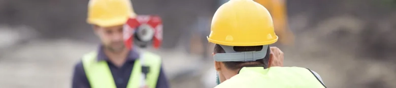 Two construction workers in safety vests and hard hats with surveying equipment in the background.