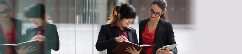 Two professional women consulting a notebook together with blurred office background.