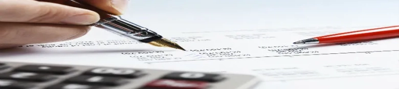 Close-up of a hand filling out a tax form with a fountain pen, with a calculator and red pencil nearby.