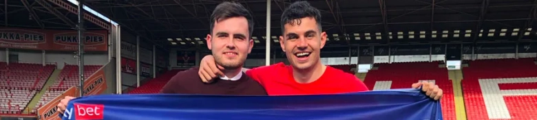 Two smiling men holding a blue banner at a football stadium with empty red seats in the background.