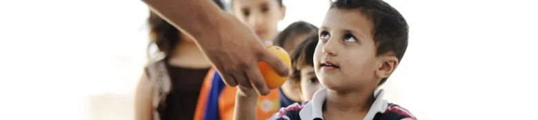 Young boy receiving an orange from an adult's hand, symbolizing food assistance in a humanitarian context.