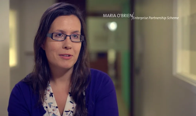 Woman in glasses and blue cardigan speaking during an interview with text overlay "MARIA O'BRIEN, Enterprise Partnership Scheme"