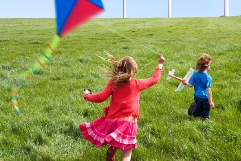 A girl running through grass with a kite with a boy in front of her