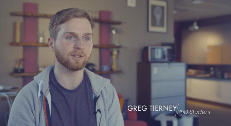 PhD student Greg Tierney in a casual hoodie, with text overlay identifying him, in a room with shelves and kitchen appliances in the background.