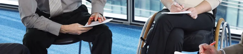 Two individuals in a professional setting taking notes, one using a wheelchair.