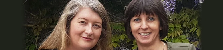 Two smiling women posing together outdoors with greenery in the background.