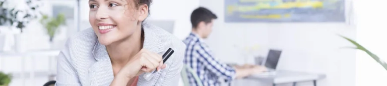 Smiling woman holding a pen with a thoughtful expression in an office environment, with a male colleague working on a laptop in the background.